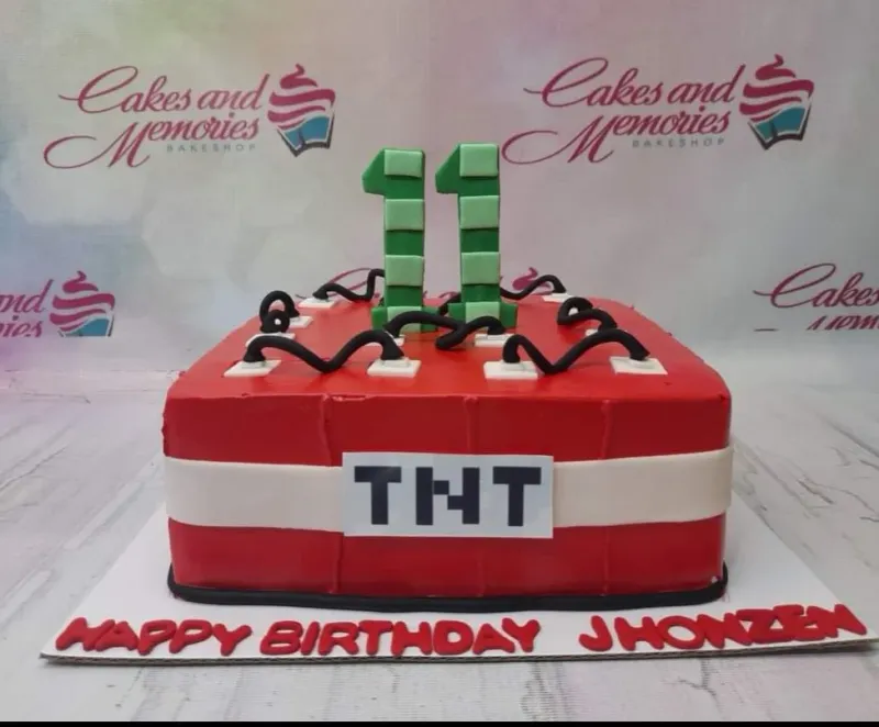 A red, green, and black square fondant cake themed around Minecraft TNT, featuring pixelated number 11 toppers and black fuse wires. A red, green, and black square fondant cake themed around Minecraft TNT, featuring pixelated number 11 toppers and black fuse wires.