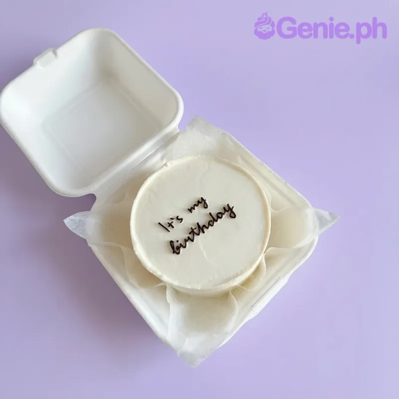 A white bento cake themed for a minimalist birthday, with black cursive icing text 'it's my birthday', presented in a white takeout box. A white bento cake themed for a minimalist birthday, with black cursive icing text 'it's my birthday', presented in a white takeout box.