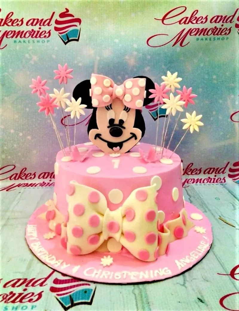 A pink and white 1-tier cake themed around Minnie Mouse, featuring a Minnie Mouse printout, a large polka dot bow, white polka dots, and fondant flowers. A pink and white 1-tier cake themed around Minnie Mouse, featuring a Minnie Mouse printout, a large polka dot bow, white polka dots, and fondant flowers.