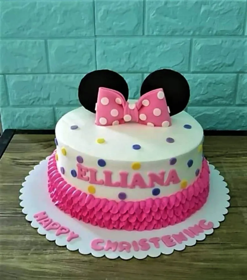 A white and pink 1-tier cake themed around Minnie Mouse, featuring black ears, a pink bow, pink ruffles, and colorful polka dots. A white and pink 1-tier cake themed around Minnie Mouse, featuring black ears, a pink bow, pink ruffles, and colorful polka dots.