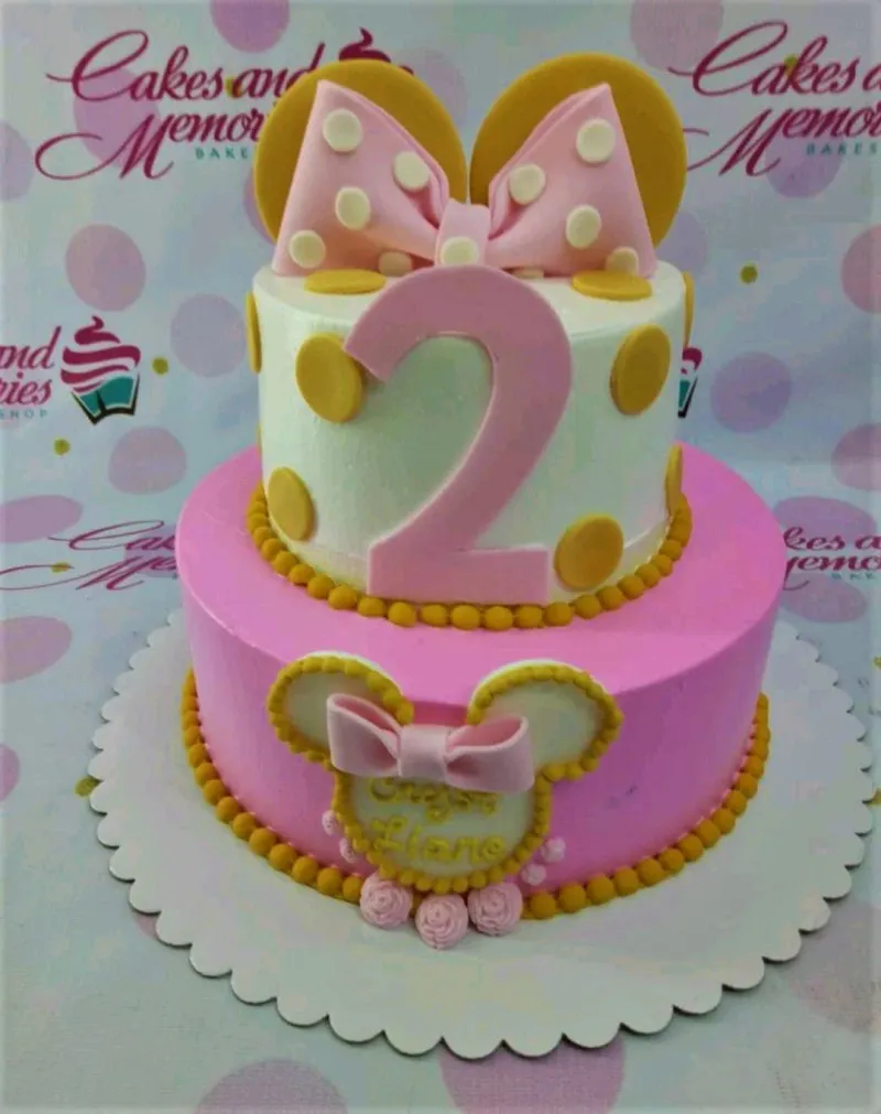 A pink and white two-tier cake themed around Minnie Mouse, featuring a pink number two, yellow ears, and a pink polka dot bow, with yellow polka dots. A pink and white two-tier cake themed around Minnie Mouse, featuring a pink number two, yellow ears, and a pink polka dot bow, with yellow polka dots.