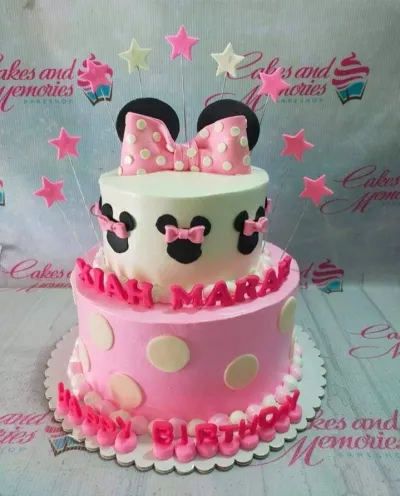 A pink and white two-tier cake themed around Minnie Mouse, featuring a large pink polka dot bow, black circular ears, and star toppers. A pink and white two-tier cake themed around Minnie Mouse, featuring a large pink polka dot bow, black circular ears, and star toppers.