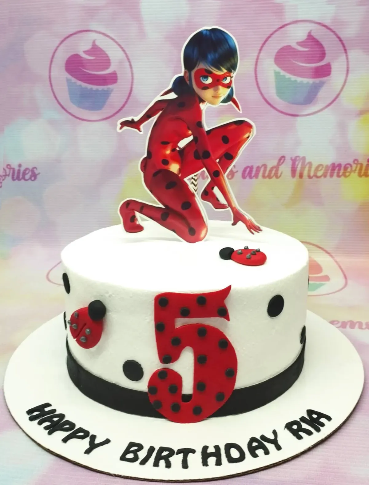 A white, red, and black 1-tier Miraculous Ladybug cake, featuring a character topper, a red number five, and black dot and ladybug accents. A white, red, and black 1-tier Miraculous Ladybug cake, featuring a character topper, a red number five, and black dot and ladybug accents.