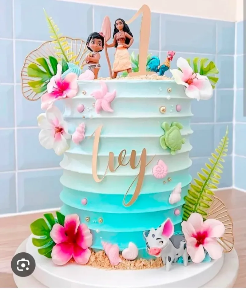 A sky blue gradient 1-tier cake with Moana character figures, tropical flowers, sea shells, and a gold name topper A sky blue gradient 1-tier cake with Moana character figures, tropical flowers, sea shells, and a gold name topper