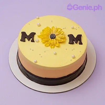 Yellow round cake for Mom featuring a large yellow icing sunflower, chocolate letters, and white pearl sprinkles on a pink background. Yellow round cake for Mom featuring a large yellow icing sunflower, chocolate letters, and white pearl sprinkles on a pink background.
