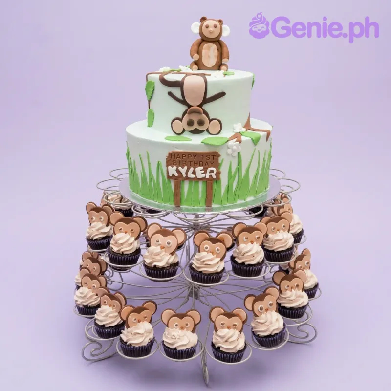 A light blue two-tier Monkey Jungle cake with fondant monkeys, green grass icing, and 'HAPPY 1ST BIRTHDAY KYLER' text. A light blue two-tier Monkey Jungle cake with fondant monkeys, green grass icing, and 'HAPPY 1ST BIRTHDAY KYLER' text.