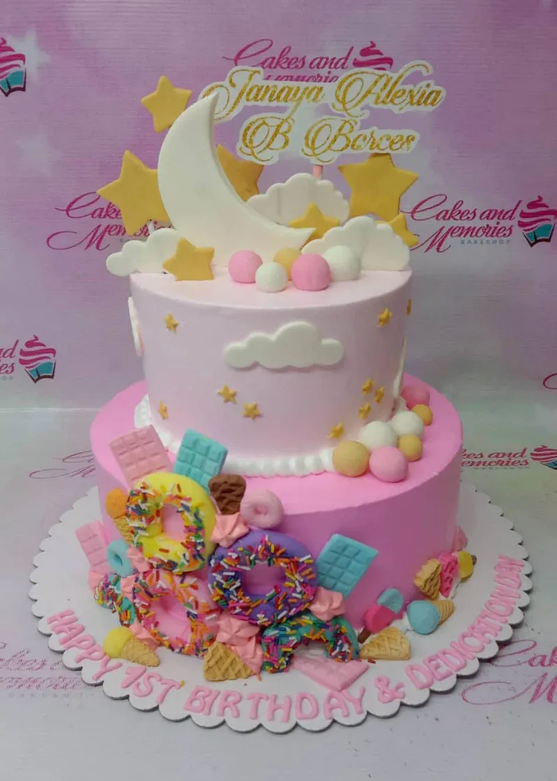 A pink two-tier cake themed Moon and Stars, featuring a white crescent moon, yellow stars, clouds, and colorful fondant treats. A pink two-tier cake themed Moon and Stars, featuring a white crescent moon, yellow stars, clouds, and colorful fondant treats.