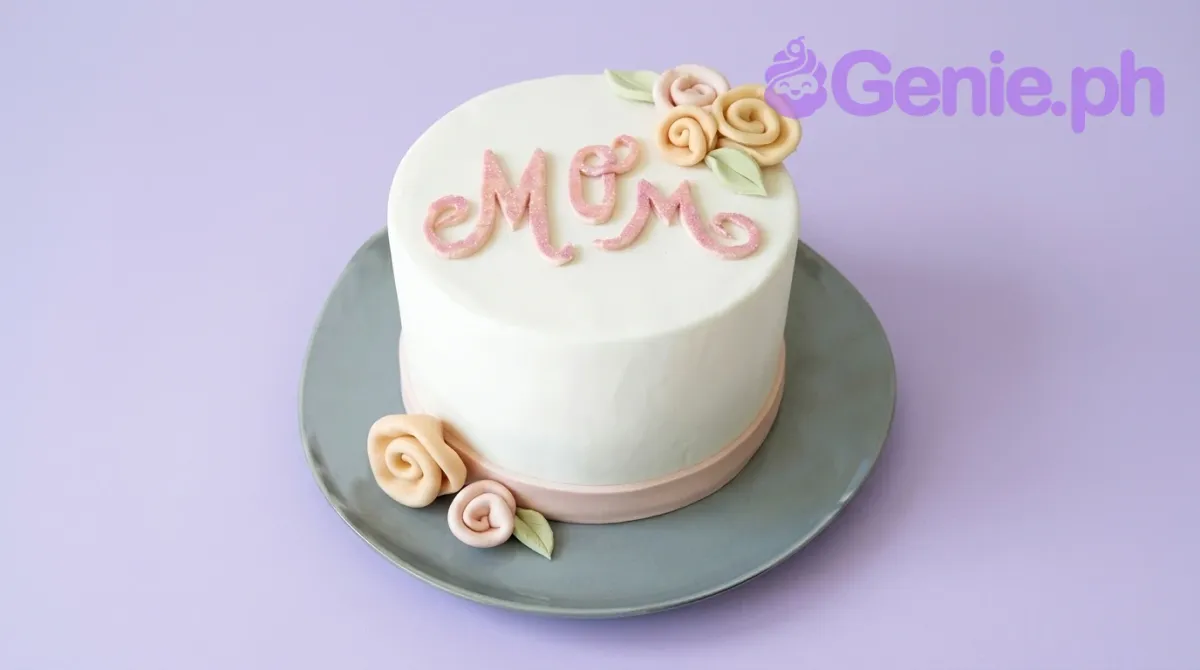 A white fondant 1-tier cake themed for Mother's Day, with pink and peach gumpaste roses and a glittery pink 'Mom' topper. A white fondant 1-tier cake themed for Mother's Day, with pink and peach gumpaste roses and a glittery pink 'Mom' topper.