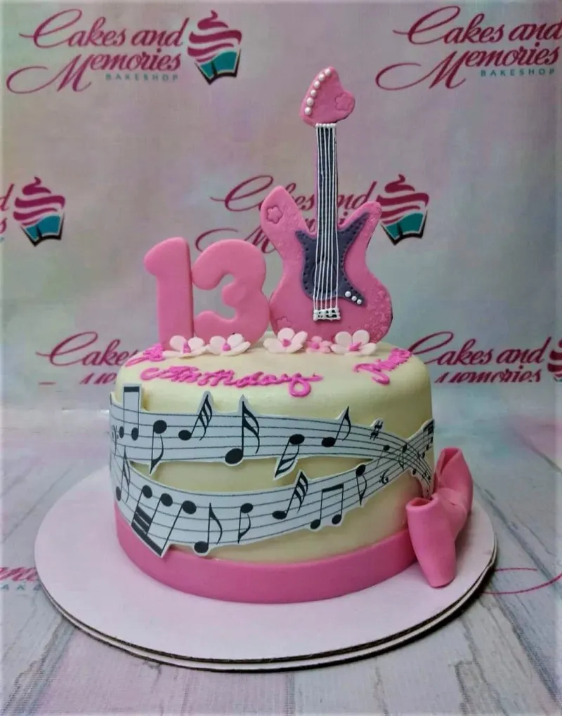 A pink and white 1-tier fondant cake themed around music, featuring a pink fondant guitar, number 13, and musical note printouts. A pink and white 1-tier fondant cake themed around music, featuring a pink fondant guitar, number 13, and musical note printouts.