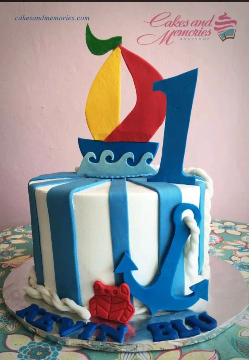 A blue and white striped 1 Tier Fondant cake, themed Nautical Sailboat, featuring a sailboat topper, a blue number one, and anchor, red crab, and rope accents. A blue and white striped 1 Tier Fondant cake, themed Nautical Sailboat, featuring a sailboat topper, a blue number one, and anchor, red crab, and rope accents.