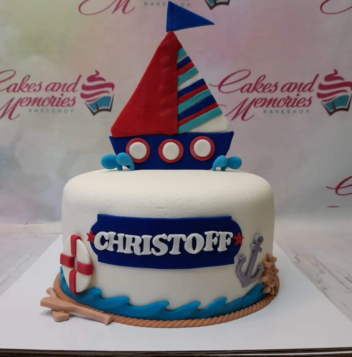 A white 1-tier fondant cake themed Nautical Sailboat, featuring a red and blue sailboat, lifebuoy, silver anchor, blue waves, and a name plaque. A white 1-tier fondant cake themed Nautical Sailboat, featuring a red and blue sailboat, lifebuoy, silver anchor, blue waves, and a name plaque.