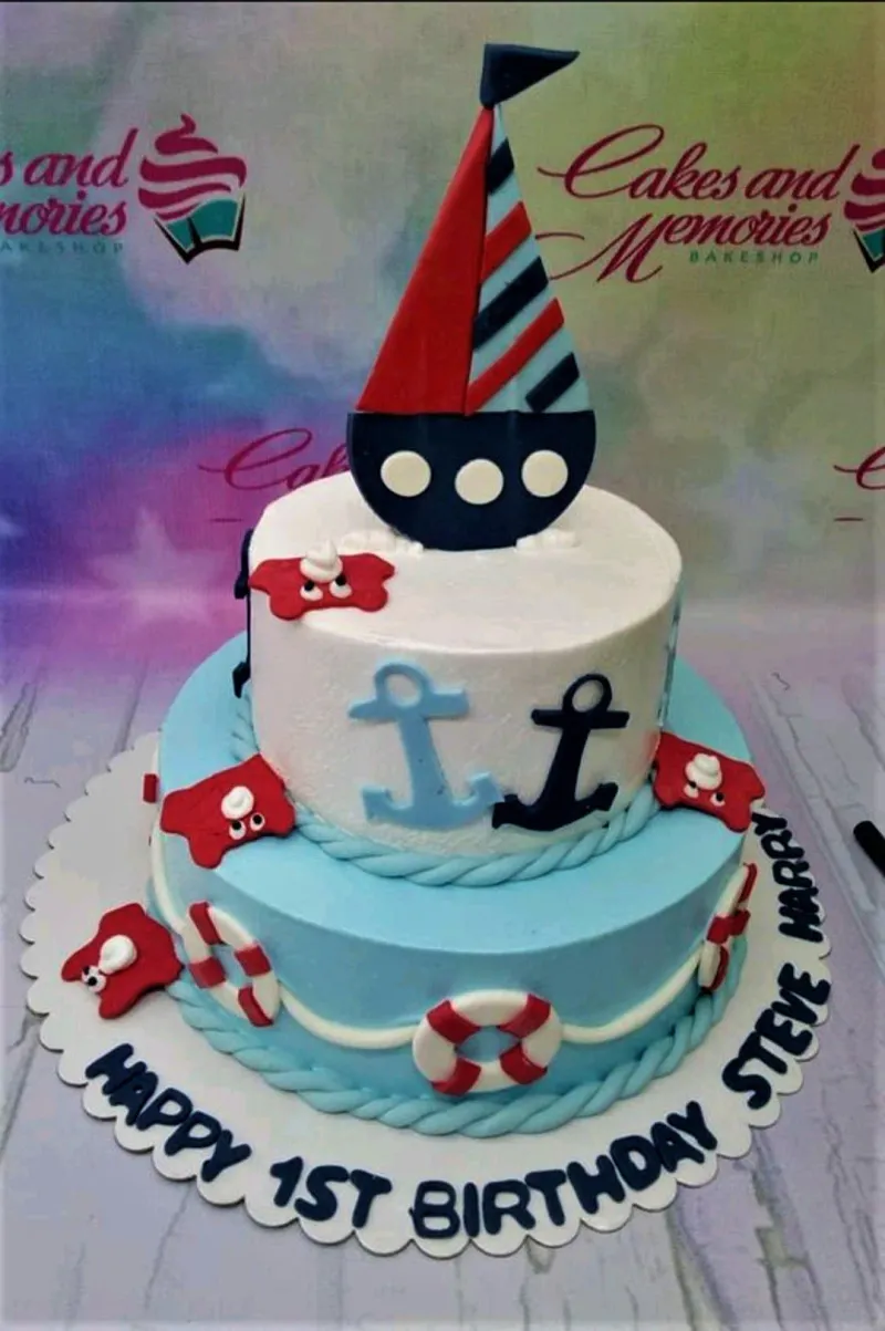 A light blue and white two-tier Nautical cake, featuring a large sailboat topper, anchors, and lifebuoys. A light blue and white two-tier Nautical cake, featuring a large sailboat topper, anchors, and lifebuoys.