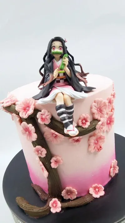 A pink 1 tier cake featuring a Nezuko figure sitting on a brown edible tree branch, surrounded by pink cherry blossom flowers. A pink 1 tier cake featuring a Nezuko figure sitting on a brown edible tree branch, surrounded by pink cherry blossom flowers.