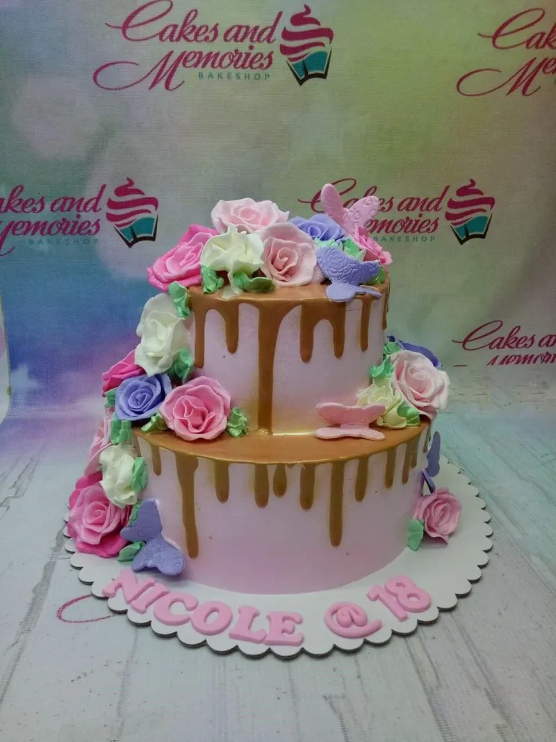 A pink two-tier cake themed for an 18th birthday, featuring edible pink and purple roses, butterflies, and gold drip. A pink two-tier cake themed for an 18th birthday, featuring edible pink and purple roses, butterflies, and gold drip.