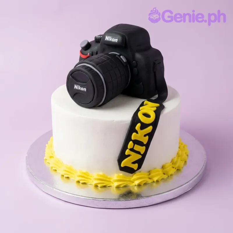 A white 1-tier cake themed around Nikon Camera, featuring a large black fondant camera topper and a yellow icing border. A white 1-tier cake themed around Nikon Camera, featuring a large black fondant camera topper and a yellow icing border.