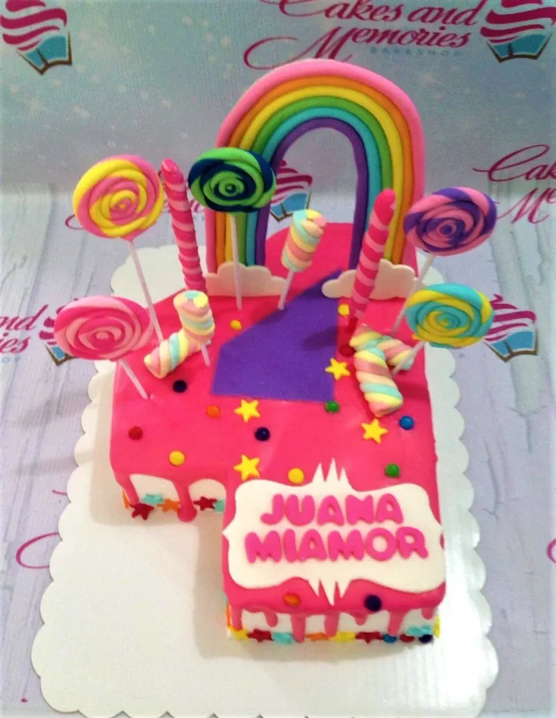 A pink number 4 shaped cake themed around rainbow, featuring a multi-colored rainbow, lollipops, marshmallows, and "JUANA MIAMOR" text. A pink number 4 shaped cake themed around rainbow, featuring a multi-colored rainbow, lollipops, marshmallows, and "JUANA MIAMOR" text.