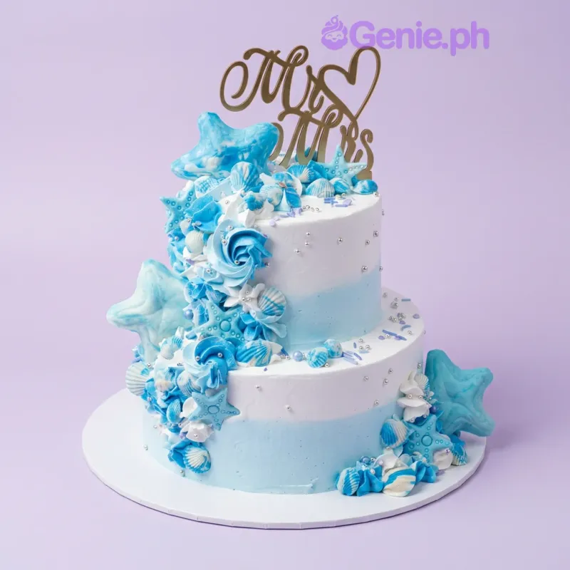 A white and blue two-tier cake themed around an Ocean Wedding, featuring a gold Mr & Mrs topper and fondant starfish, seashells, and silver dragees. A white and blue two-tier cake themed around an Ocean Wedding, featuring a gold Mr & Mrs topper and fondant starfish, seashells, and silver dragees.