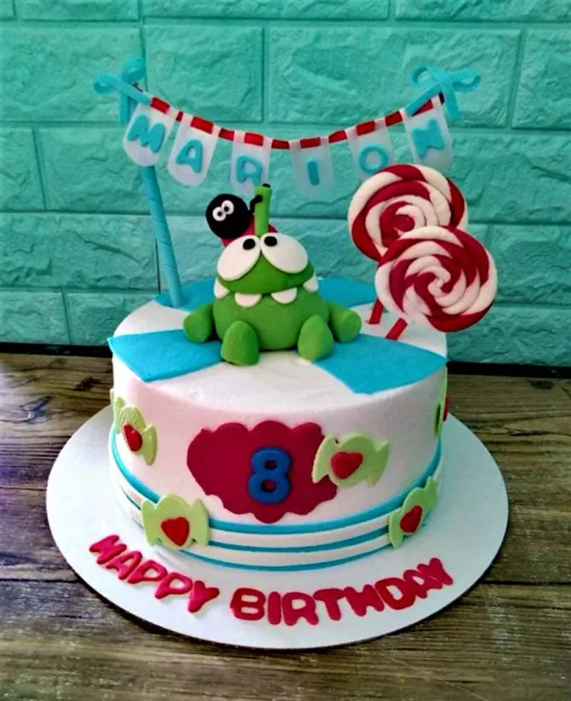 Om Nom Cut the Rope themed birthday cake with green fondant character, lollipops, and a name banner. Om Nom Cut the Rope themed birthday cake with green fondant character, lollipops, and a name banner.