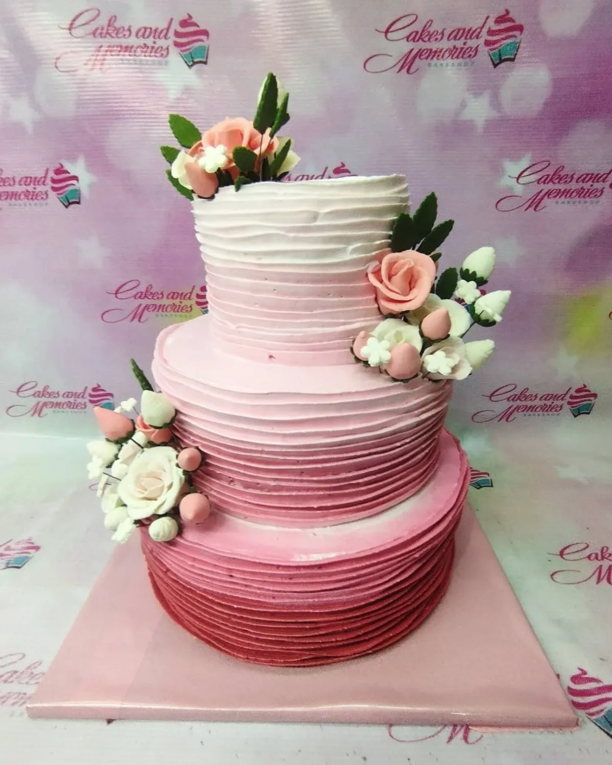 A three-tier ombre pink floral cake with horizontal icing ridges, featuring peach and white gumpaste roses and buds. A three-tier ombre pink floral cake with horizontal icing ridges, featuring peach and white gumpaste roses and buds.