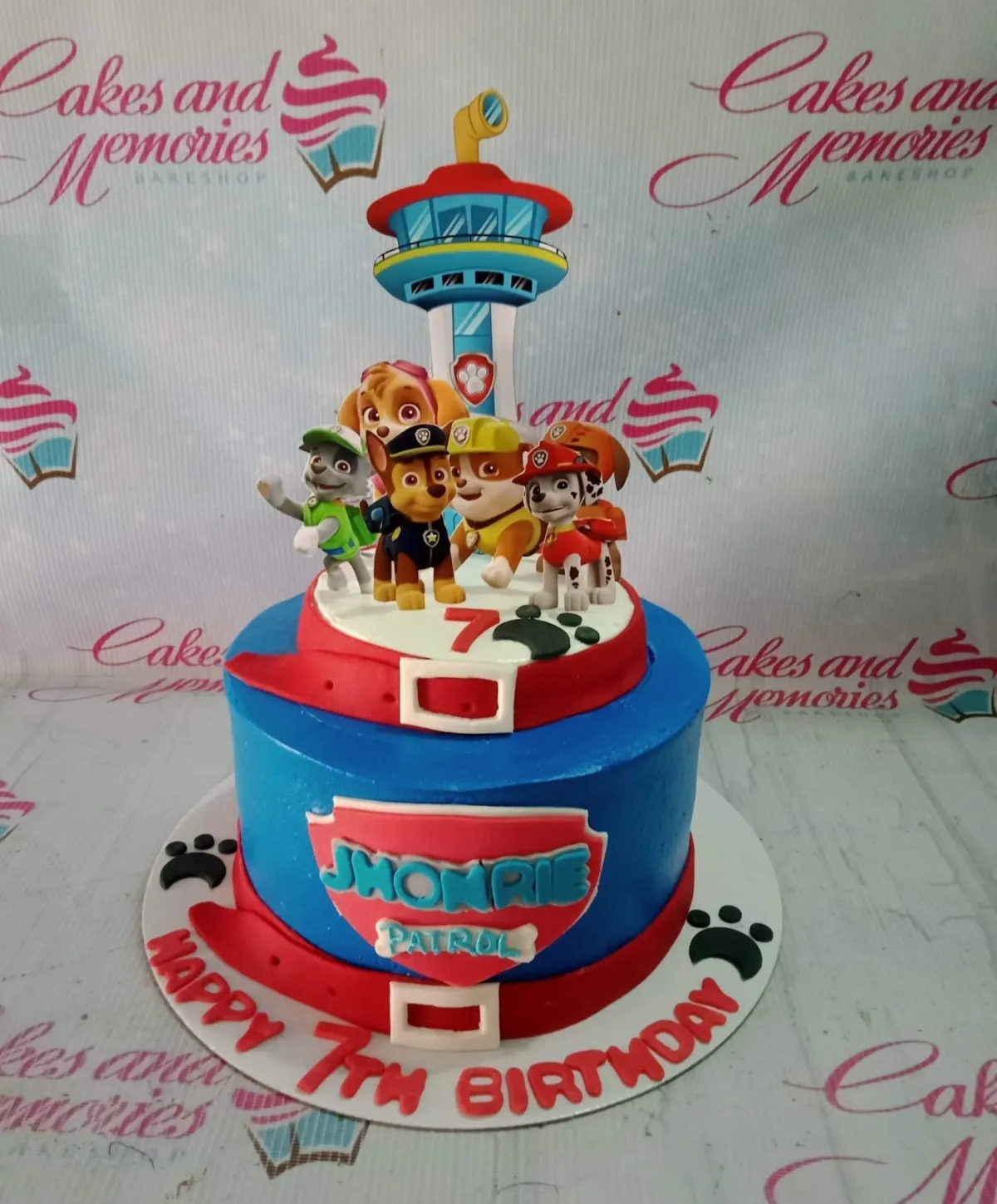 A blue 1-tier Paw Patrol cake with a lookout tower printout, character cutouts, red fondant belts, and paw prints. A blue 1-tier Paw Patrol cake with a lookout tower printout, character cutouts, red fondant belts, and paw prints.