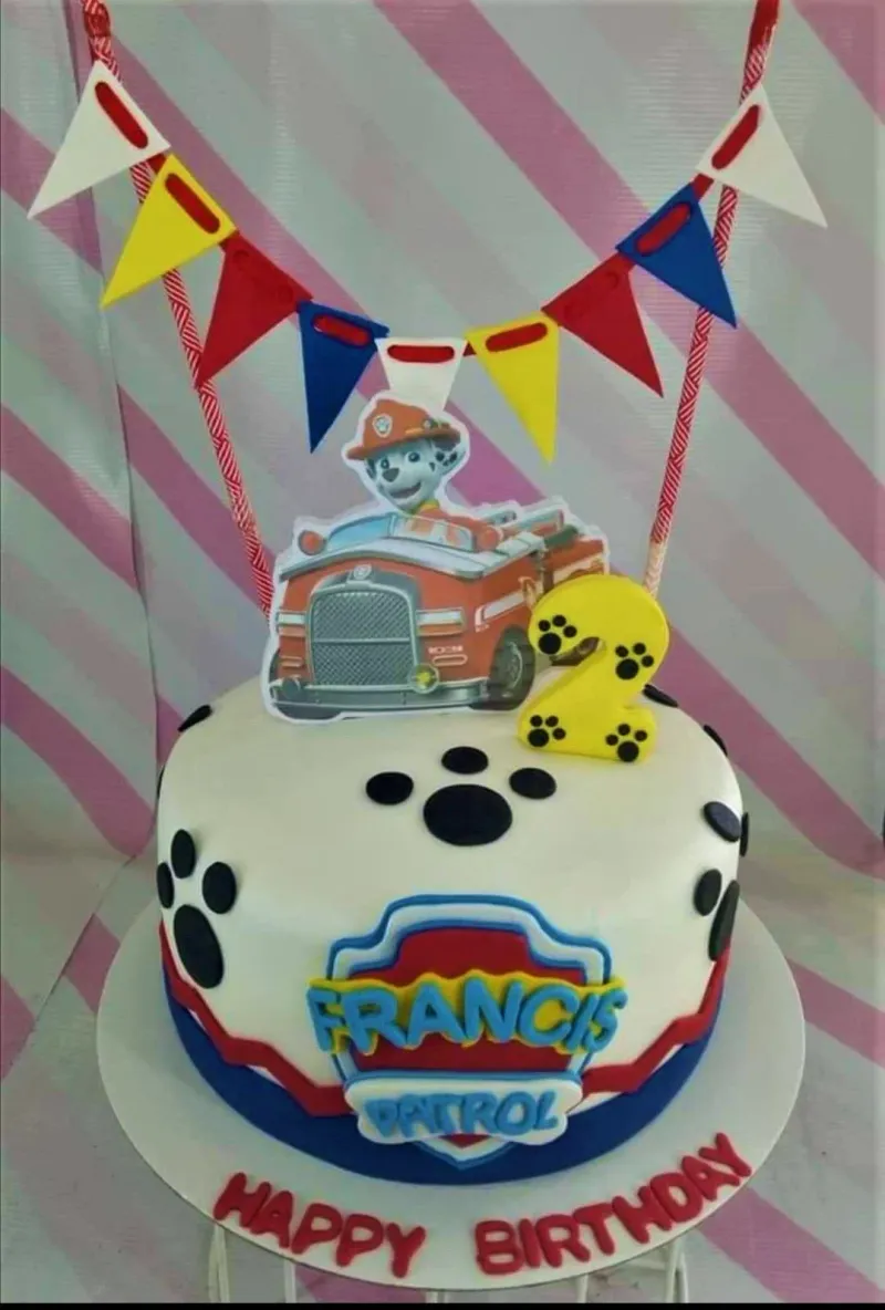 A white 1-tier fondant cake themed Paw Patrol, featuring Marshall in a fire truck printout, a yellow number 2, and paw print decorations. A white 1-tier fondant cake themed Paw Patrol, featuring Marshall in a fire truck printout, a yellow number 2, and paw print decorations.