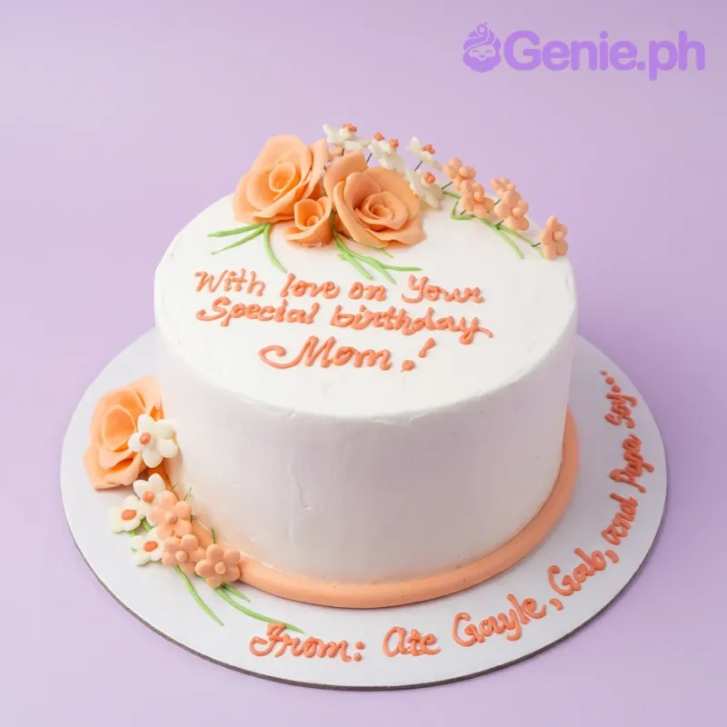 A white 1-tier round Mom cake, featuring peach and white flowers, orange piped text, and an orange icing border. A white 1-tier round Mom cake, featuring peach and white flowers, orange piped text, and an orange icing border.