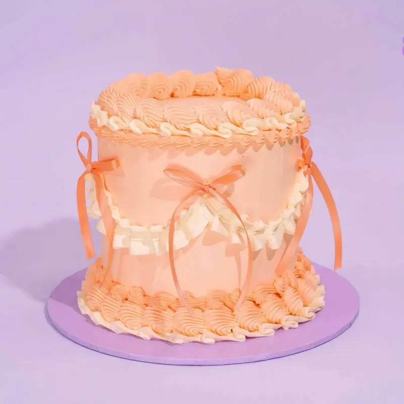 Peach 1-tier cake with piped icing ruffles and satin ribbon bows on a pink cake board Peach 1-tier cake with piped icing ruffles and satin ribbon bows on a pink cake board