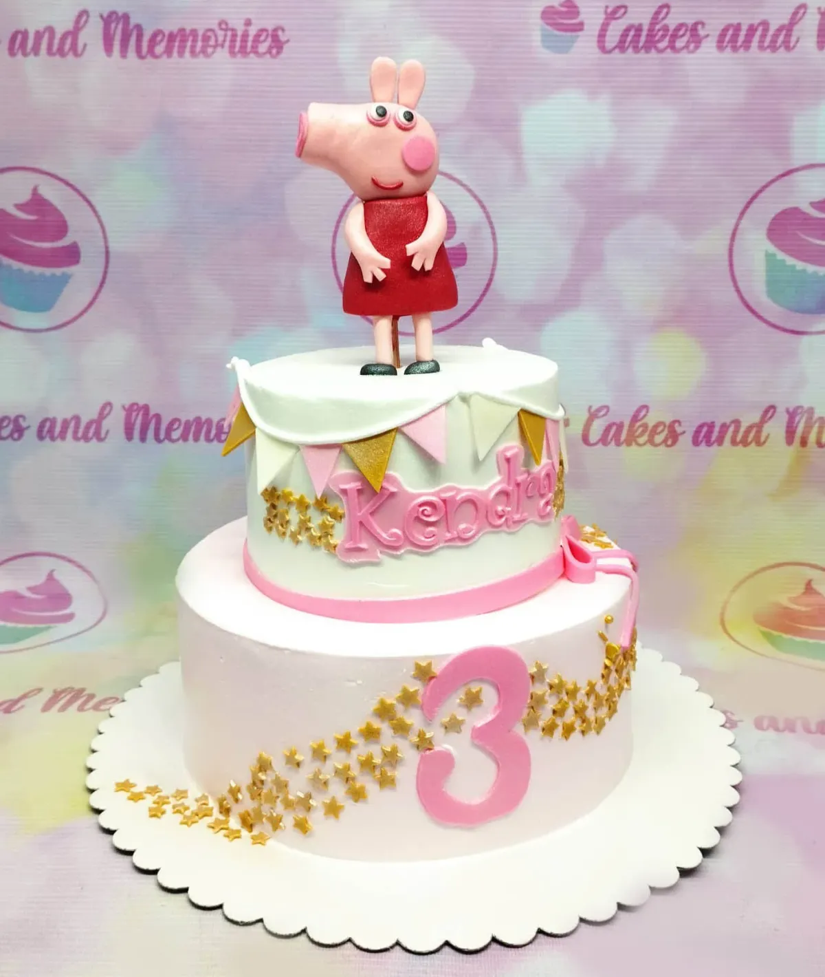 A white and light pink 2 Tier Fondant cake themed around Peppa Pig, featuring a 3D Peppa Pig figure, gold stars, pink bunting, and gumpaste letters 'Kendra 3'. A white and light pink 2 Tier Fondant cake themed around Peppa Pig, featuring a 3D Peppa Pig figure, gold stars, pink bunting, and gumpaste letters 'Kendra 3'.