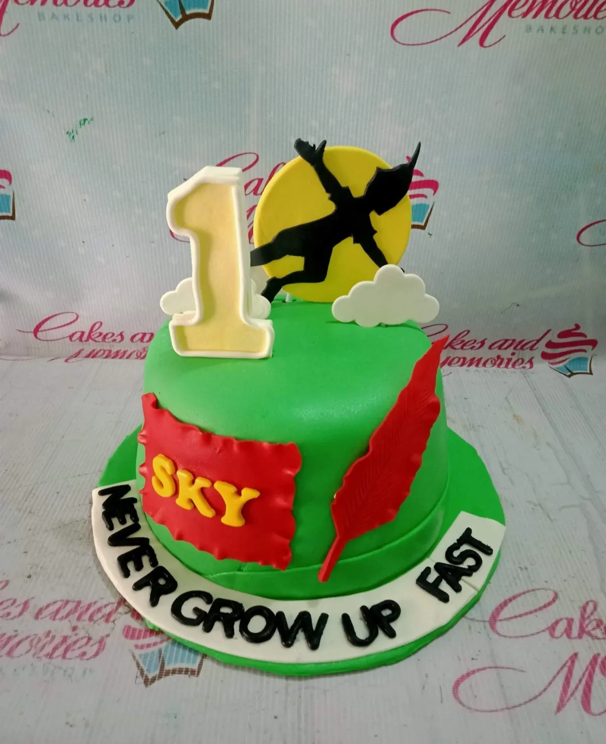 A green 1-tier Peter Pan cake, featuring a Peter Pan silhouette, yellow moon, number one, and a red feather. A green 1-tier Peter Pan cake, featuring a Peter Pan silhouette, yellow moon, number one, and a red feather.