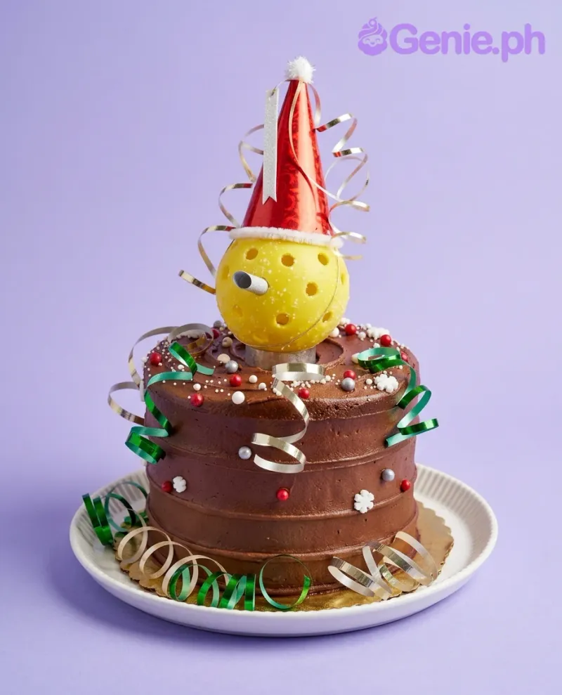 A brown 1 Tier Pickleball cake, featuring a yellow pickleball topper with a red party hat, silver and green ribbons. A brown 1 Tier Pickleball cake, featuring a yellow pickleball topper with a red party hat, silver and green ribbons.