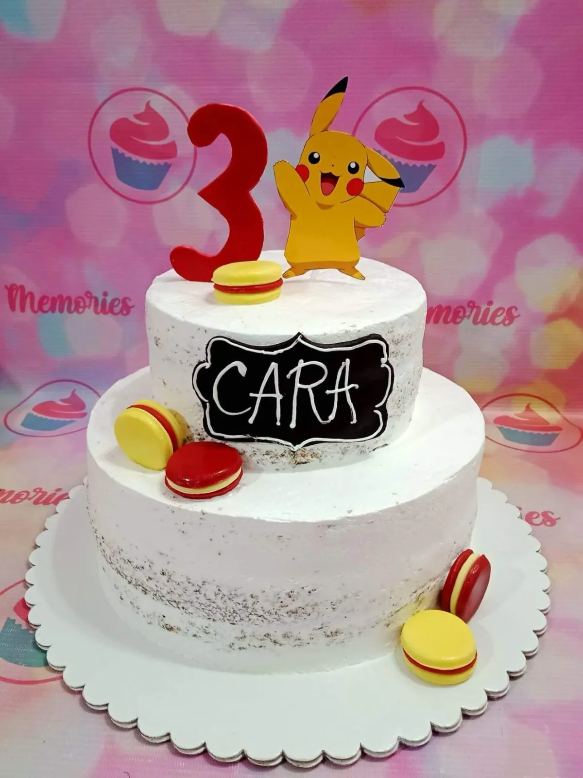 A white two-tier Pikachu-themed cake with a Pikachu printout, red number three topper, yellow and red macarons, and a black name plaque. A white two-tier Pikachu-themed cake with a Pikachu printout, red number three topper, yellow and red macarons, and a black name plaque.
