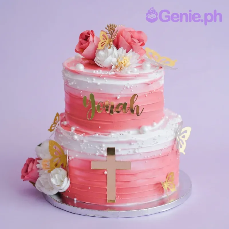 A pink and white gradient two-tier baptism cake, with a gold cross, gold name, pink roses, white flowers, gold butterflies, and pearls. A pink and white gradient two-tier baptism cake, with a gold cross, gold name, pink roses, white flowers, gold butterflies, and pearls.