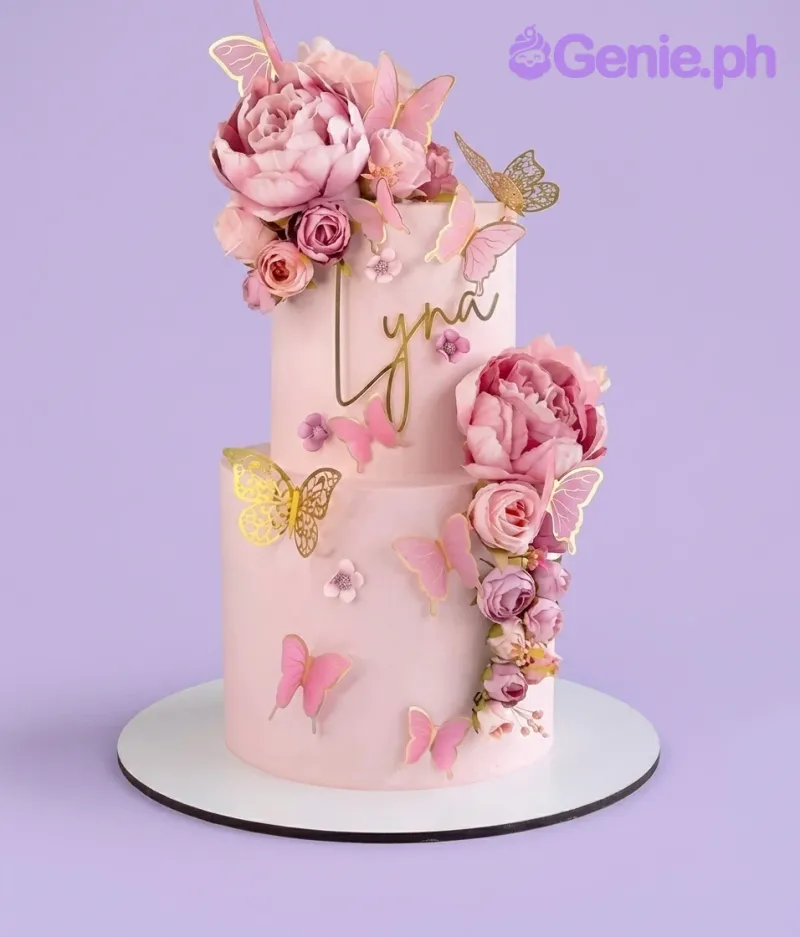 A light pink 2-tier cake with a Pink Butterfly Floral theme, featuring a gold name cutout, pink peonies, roses, and various pink and gold butterflies. A light pink 2-tier cake with a Pink Butterfly Floral theme, featuring a gold name cutout, pink peonies, roses, and various pink and gold butterflies.