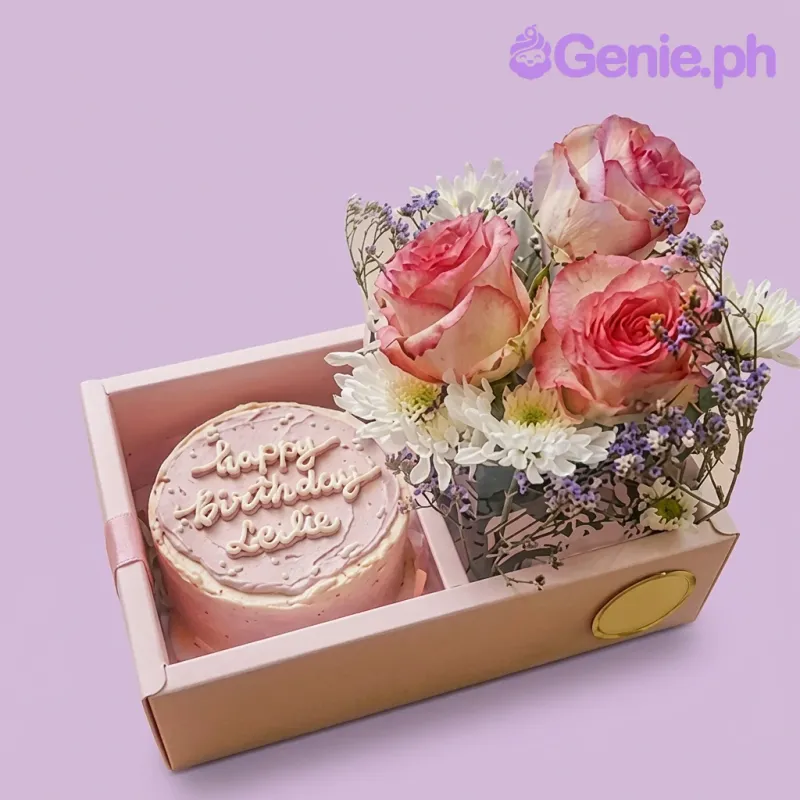 A pink 1 tier cake with 'Happy Birthday Leslie' text, fresh pink roses, and white filler flowers in a clear gift box. A pink 1 tier cake with 'Happy Birthday Leslie' text, fresh pink roses, and white filler flowers in a clear gift box.