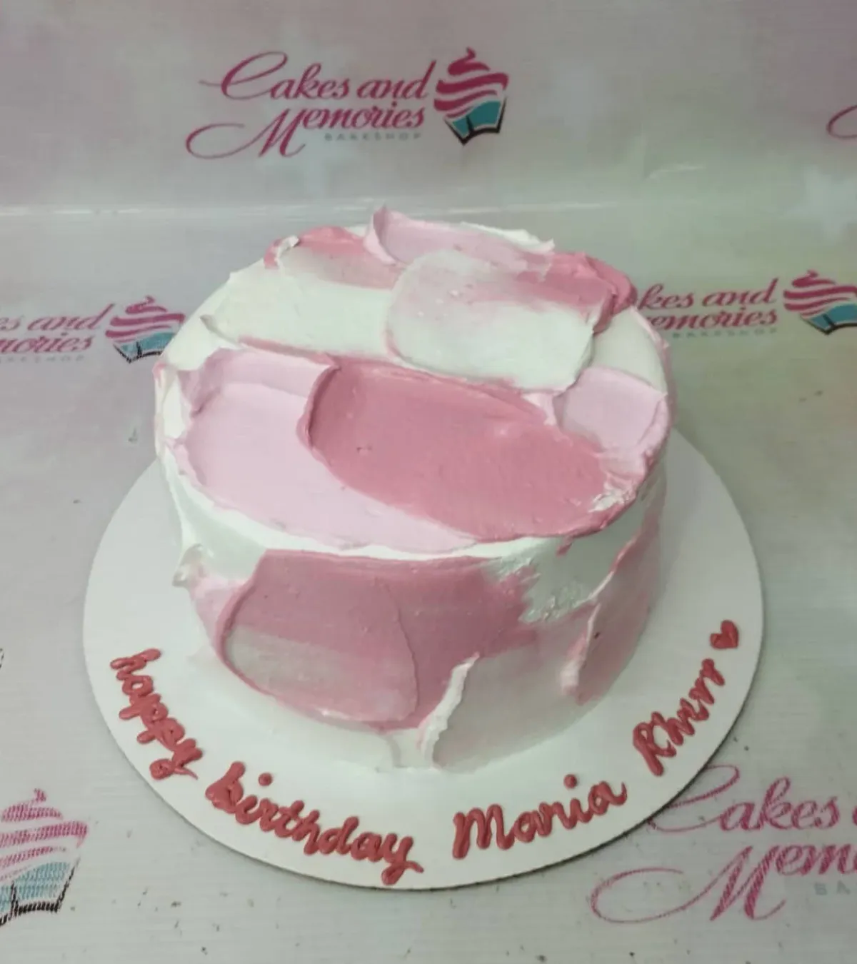 A white 1-tier cake with pink and light pink palette knife icing strokes, a small pink heart, and 'happy birthday Maria Kherr' text. A white 1-tier cake with pink and light pink palette knife icing strokes, a small pink heart, and 'happy birthday Maria Kherr' text.