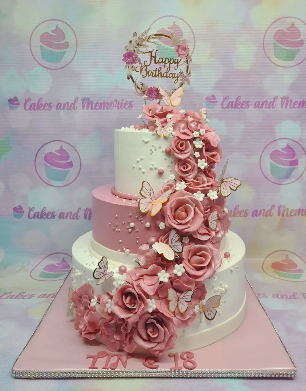 A pink and white 3-tier debut cake, with a gold Happy Birthday topper, cascading pink roses, and pink butterflies. A pink and white 3-tier debut cake, with a gold Happy Birthday topper, cascading pink roses, and pink butterflies.