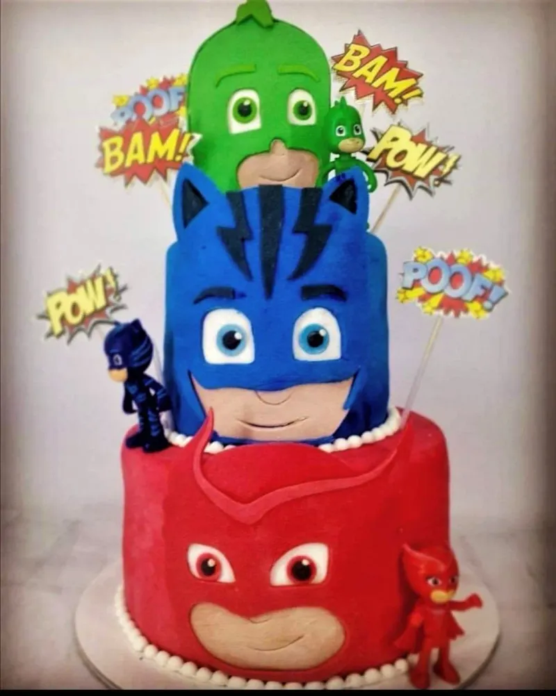 A PJ Masks themed 3-tier fondant cake featuring Gekko, Catboy, and Owlette character faces with comic book action bubbles. A PJ Masks themed 3-tier fondant cake featuring Gekko, Catboy, and Owlette character faces with comic book action bubbles.