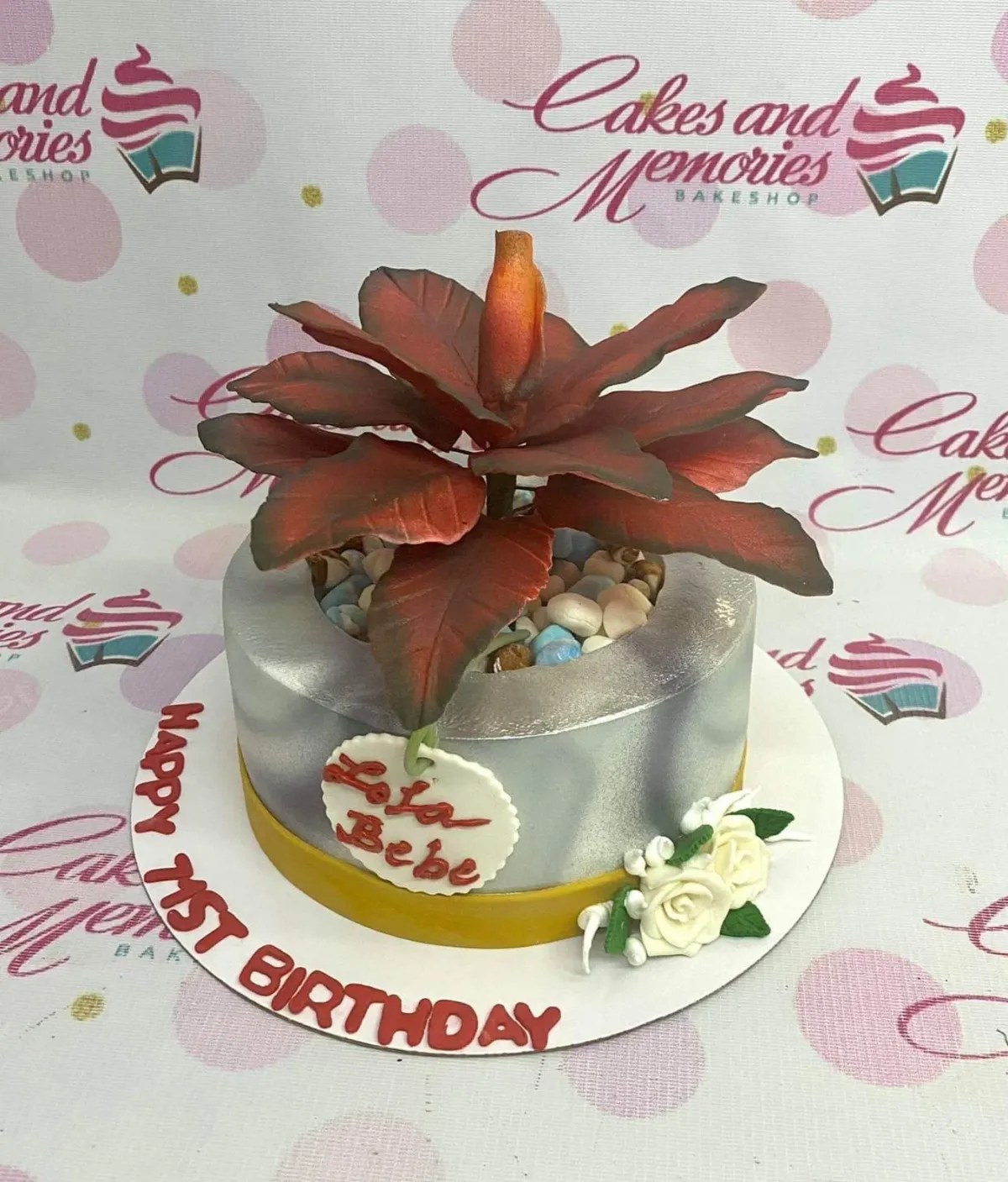 A silver metallic 1 Tier Fondant cake, shaped like a plant pot, featuring a reddish-brown gumpaste plant and white roses. A silver metallic 1 Tier Fondant cake, shaped like a plant pot, featuring a reddish-brown gumpaste plant and white roses.