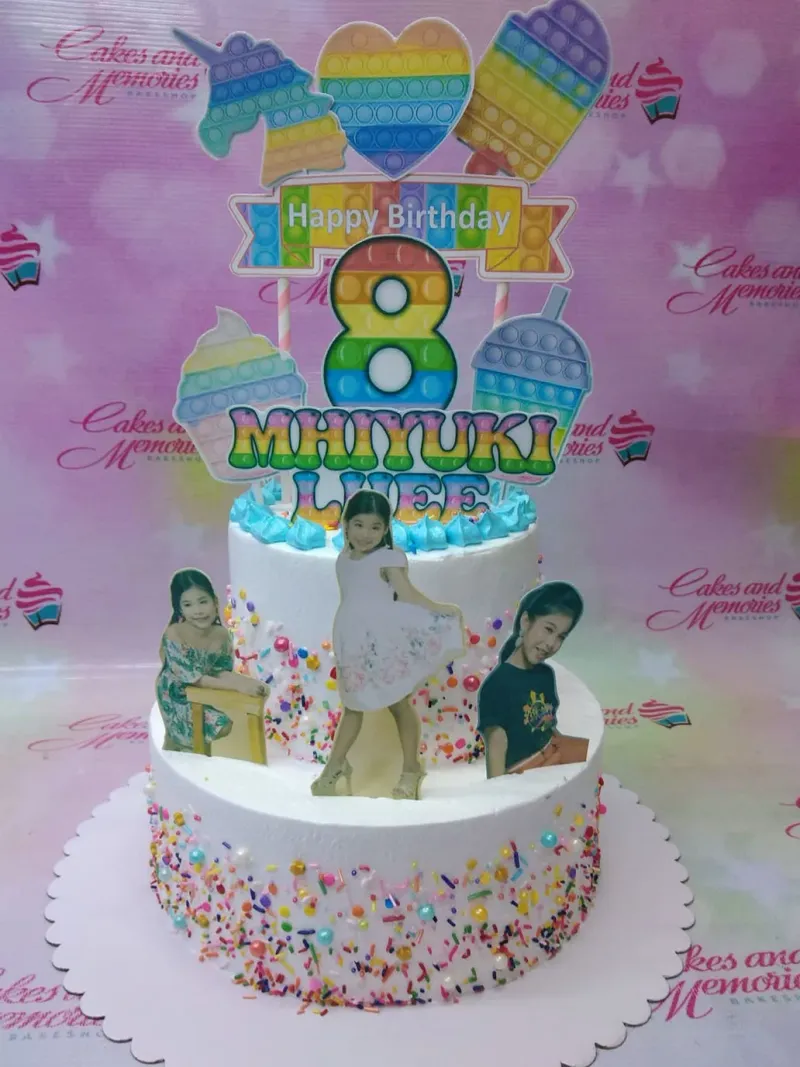 A white two-tier Pop-It themed cake, featuring a rainbow pop-it number 8, a girl's photo, and pop-it character printouts, with multi-colored sprinkles. A white two-tier Pop-It themed cake, featuring a rainbow pop-it number 8, a girl's photo, and pop-it character printouts, with multi-colored sprinkles.