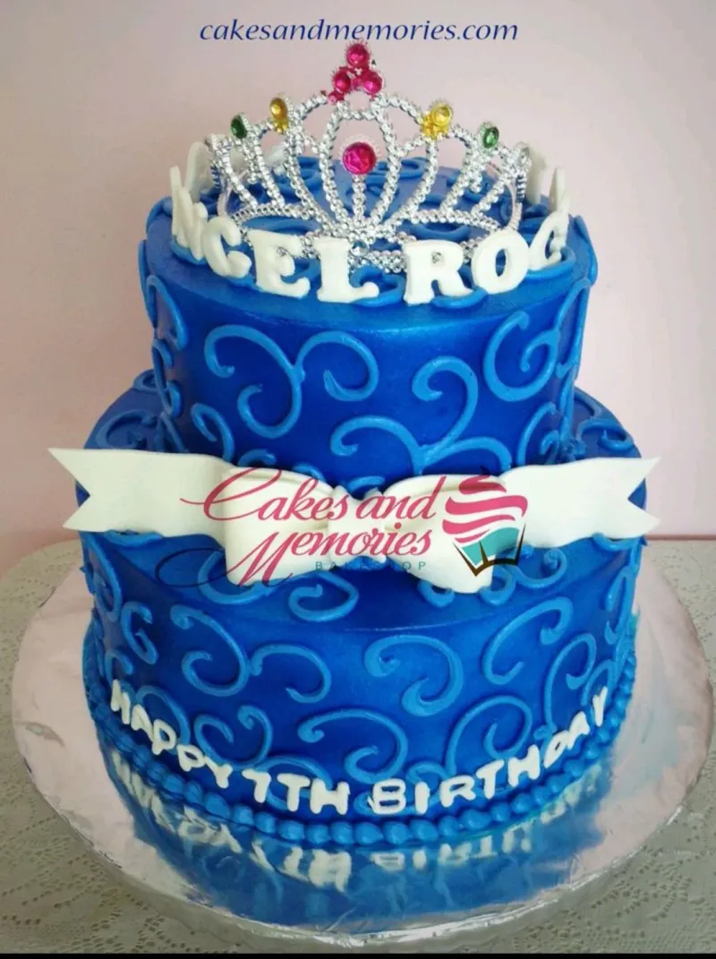 Two-tier royal blue fondant cake with intricate light blue icing swirls, a white fondant bow, and a silver rhinestone tiara on top. Two-tier royal blue fondant cake with intricate light blue icing swirls, a white fondant bow, and a silver rhinestone tiara on top.