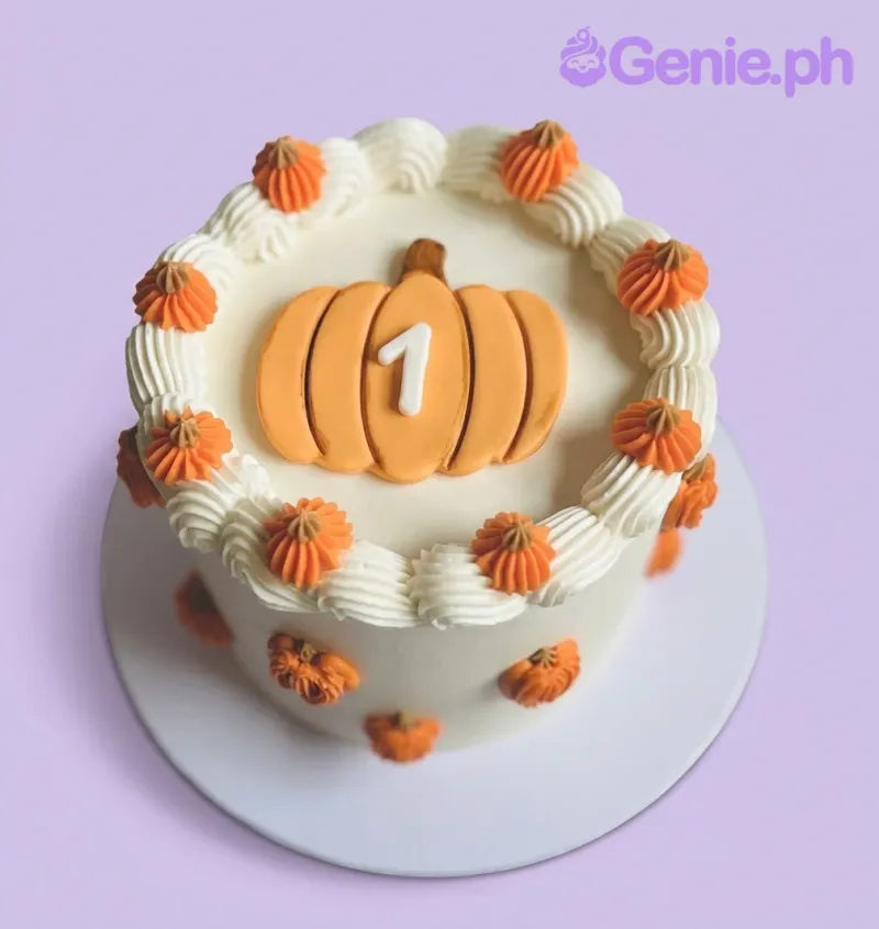 A white 1-tier cake themed Pumpkin First Birthday, with a large orange fondant pumpkin topper and orange icing rosettes. A white 1-tier cake themed Pumpkin First Birthday, with a large orange fondant pumpkin topper and orange icing rosettes.