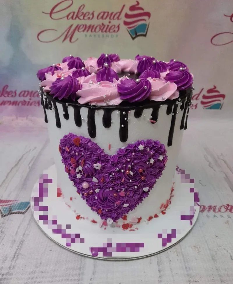 A white 1-tier cake with chocolate drip, purple and pink rosettes, a large purple textured heart, silver dragees, and heart sprinkles. A white 1-tier cake with chocolate drip, purple and pink rosettes, a large purple textured heart, silver dragees, and heart sprinkles.