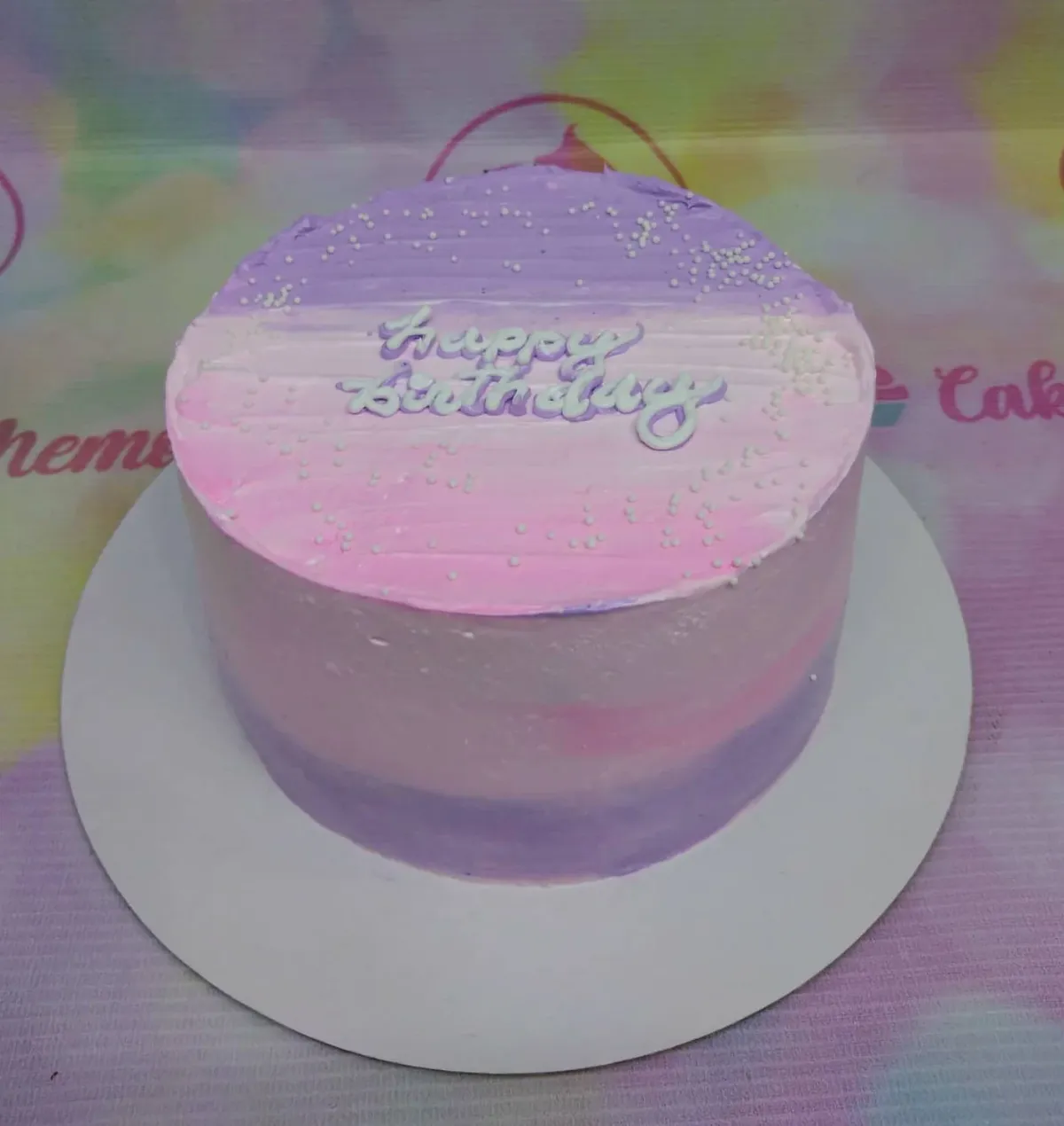 A purple and pink ombre 1-tier cake themed around a birthday, featuring a 'Happy Birthday' text topper and white sprinkles. A purple and pink ombre 1-tier cake themed around a birthday, featuring a 'Happy Birthday' text topper and white sprinkles.