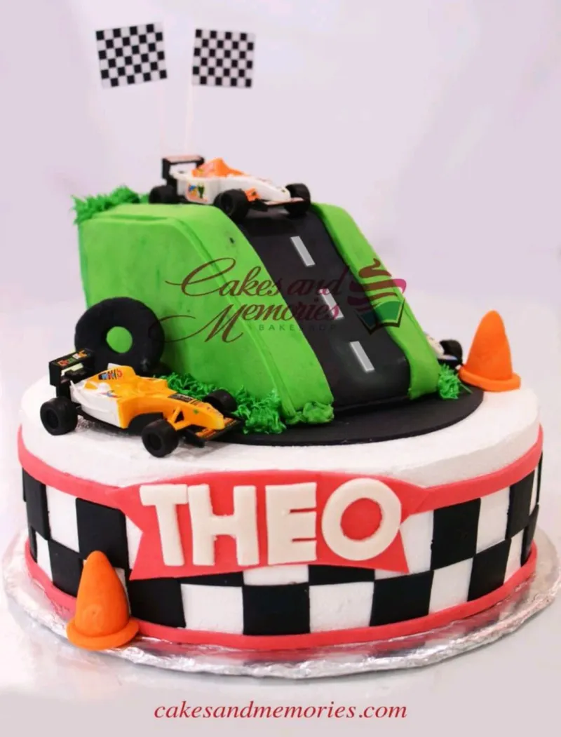 Racing car themed cake with a green hill, black road, and toy race cars on a checkered base. Racing car themed cake with a green hill, black road, and toy race cars on a checkered base.