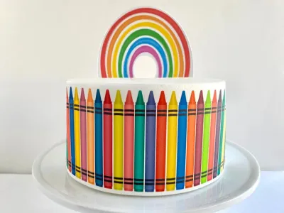 A white single tier cake themed around rainbow crayons, featuring a colorful rainbow topper and a side wrap of printed crayons. A white single tier cake themed around rainbow crayons, featuring a colorful rainbow topper and a side wrap of printed crayons.