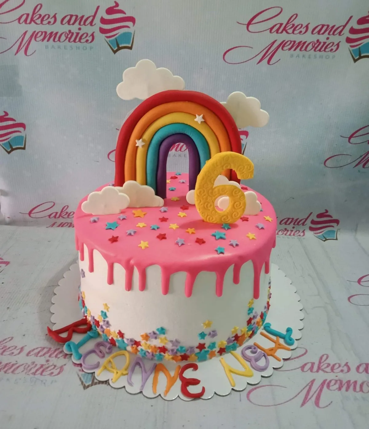 A white and pink 1-tier Rainbow cake, featuring a rainbow topper, yellow number 6, clouds, colorful star sprinkles, and 'Leanne Noel' text. A white and pink 1-tier Rainbow cake, featuring a rainbow topper, yellow number 6, clouds, colorful star sprinkles, and 'Leanne Noel' text.