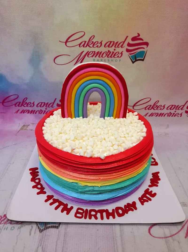 A multi-colored 1 Tier Rainbow cake, featuring a large fondant rainbow topper, horizontal icing stripes, and white marshmallow clouds. A multi-colored 1 Tier Rainbow cake, featuring a large fondant rainbow topper, horizontal icing stripes, and white marshmallow clouds.