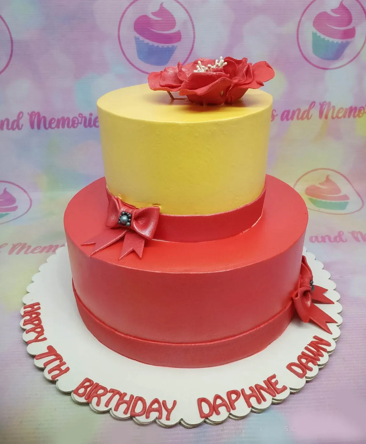 A yellow and red two-tier cake themed around Red And Yellow Floral, featuring a large red edible flower topper and red fondant bows with pearl centers. A yellow and red two-tier cake themed around Red And Yellow Floral, featuring a large red edible flower topper and red fondant bows with pearl centers.