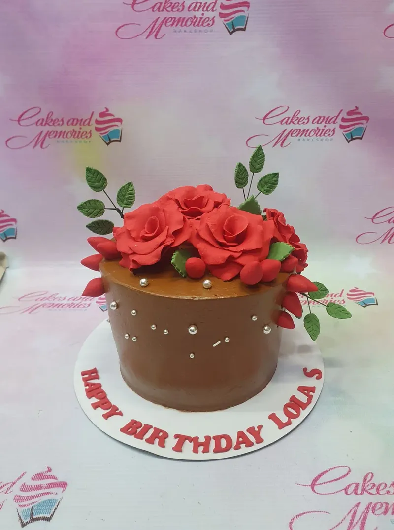 A brown 1-tier Red Rose Chocolate cake, featuring large red fondant roses, green leaves, silver dragees, and a birthday message. A brown 1-tier Red Rose Chocolate cake, featuring large red fondant roses, green leaves, silver dragees, and a birthday message.