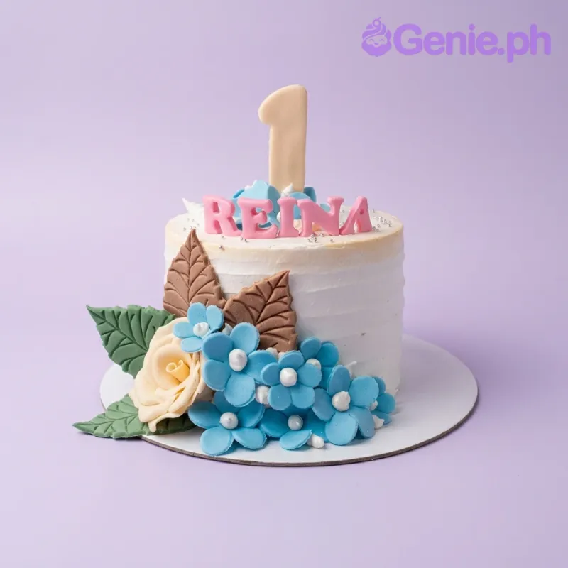 A white 1 Tier Reina Floral cake featuring a pink 'REINA' topper, a large number one, blue flowers, and green and brown leaves. A white 1 Tier Reina Floral cake featuring a pink 'REINA' topper, a large number one, blue flowers, and green and brown leaves.
